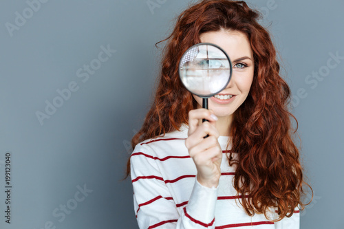 Laboratory equipment. Close up of magnifying glass being in hands of a cheerful smart red haired woman while doing a research