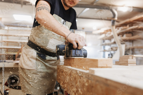 Carpenter working with electric planer on wooden plank in workshop. Craftsman makes own successful small business.