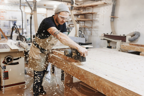 Carpenter working with electric planer on wooden plank in workshop. Craftsman makes own successful small business.