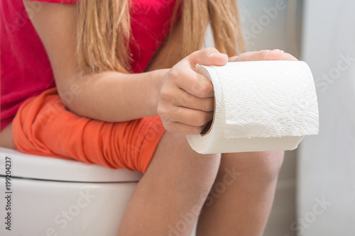 A girl is holding a roll of toilet paper in her hands, sitting on the toilet