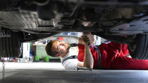 Automechaniker repariert Fahrzeug in einer Werkstatt in Arbeitskleidung am Unterboden - Closeup // Car mechanic repairs vehicle in a workshop in work clothes on the underbody