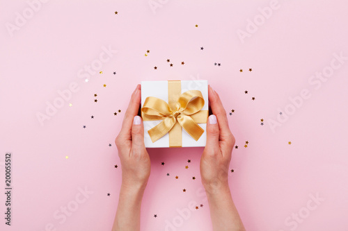 Womans hands holding gift or present box decorated confetti on pink pastel table top view. Flat lay composition for birthday or wedding.