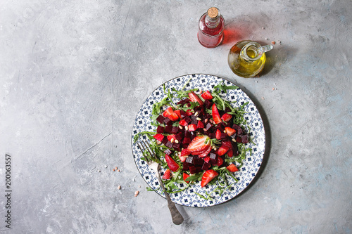 Beetroot and strawberry salad served with arugula and nuts on ceramic plate with bottles of fruit ocet and olive oil over grey texture background. Top view, copy space. Healthy eating