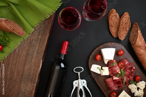 Above overhead view flat lay still life of assortment various cheese and delicatessen with traditional bread and red wine on a old wooden board on black table