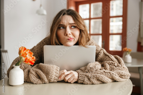 Photo of disappointed woman in sweater looking aside with suspicious gaze, while hugging laptop in coffee shop