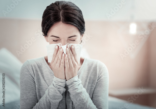 Home treatment. Ill exhausted unhappy woman sitting in the bright room holding a napkin and sneezing.