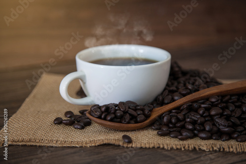 Hot coffee in white cup and coffee beans in spoon on wooden table 