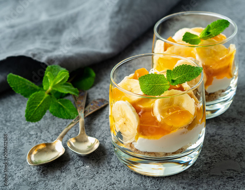 Healthy layered dessert with yogurt, banana, mango jam, cookie in glass on gray stone background, side view.