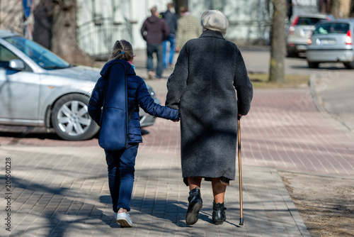 Old woman walking down the street with walking stick and a little girl goes with her. View from the back.