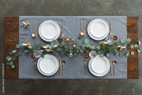 Top view of rustic table setting with eucalyptus, tarnished cutlery, wine glasses, candles and empty plates on tabletop
