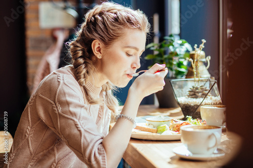 Charming woman with cute hairstyle having lunch during the rest in coffee shop, happy Caucasian female eating breakfast while relaxing in cafe during free time