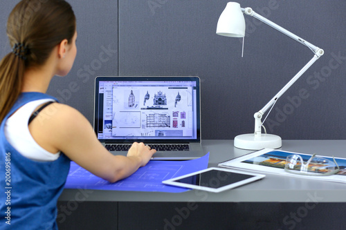 Young woman sitting at the desk with instruments, plan and laptop