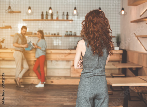 Abused young woman looking at her boyfriend cheating with young girl. She is standing in cafe with crossed hands. Focus on female back