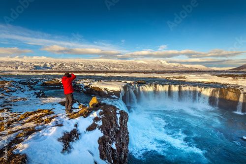 Photoghaper taking a photo at Godafoss waterfall in winter, Iceland.