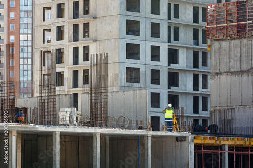 The builder in yellow overalls makes a topographic survey against the background of a concrete wall, a multi-apartment building under construction.