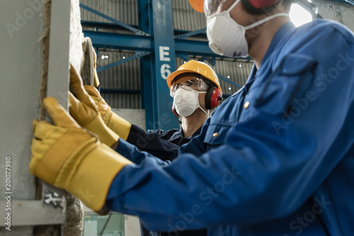 Two blue-collar workers wearing protective equipment while insulating an industrial pressure vessel