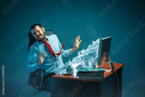 Young stressed handsome businessman working at desk in modern office shouting at laptop screen and being angry about spam