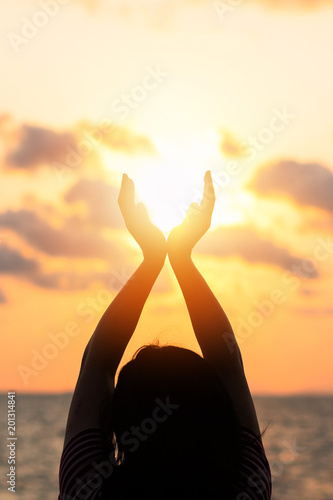 June summer sun solstice concept and silhouette of happy young woman’s hands relaxing, meditating and holding sunset against warm golden hour sky on the beach with ocean or sea background