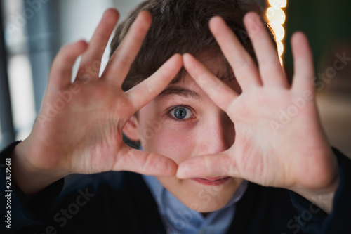 The teenage boy with blue eyes playfully looks forward through the frame from his fingers. Close up.