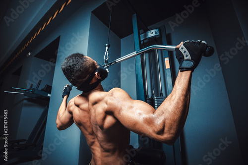 african american athletic man doing exercise in pull down machine back view. black fitness man working out lat pulldown training at gym.
