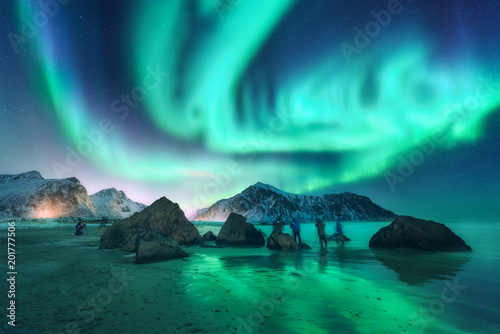 Green aurora borealis and photographers. Aurora. Northern lights in Lofoten islands, Norway. Starry sky with polar lights. Night landscape with aurora, sea, people, stones, sandy beach and mountains.