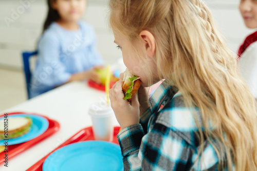 Little schoolgirl with beautiful blond hair eating tasty sandwich while sitting at table with classmates in elementary school cafeteria