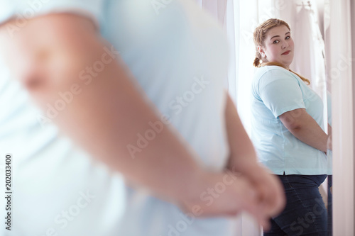 Mirror lies. Nice young woman looking disappointed while evaluating her figure in the mirror
