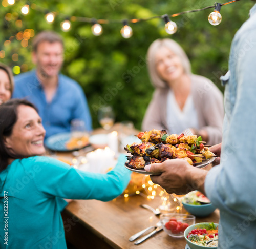  group of friends in their forties gathered around a table in the garden to share a bbq meal. A man offers chicken skewers to guests