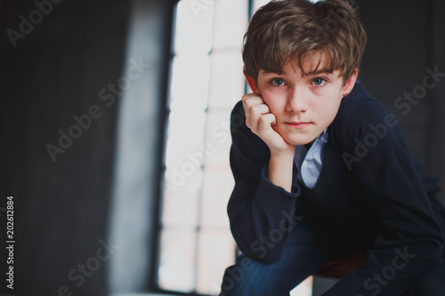 Sad brooding teen boy with blue eyes in blue casual wear sits on a chair in a dark loft style room and looks right into the camera. In the background large window.