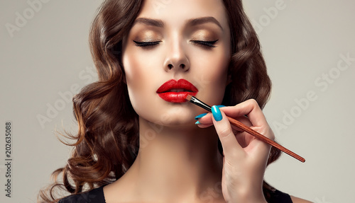 Makeup artist applies red lipstick . Beautiful woman face. Hand of make-up master, painting lips of young beauty model girl . Make up in process