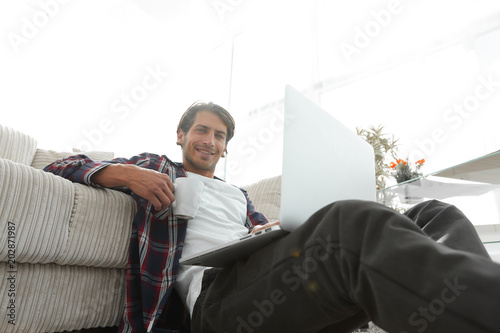 young man with laptop holding a cup sitting on the floor near the sofa