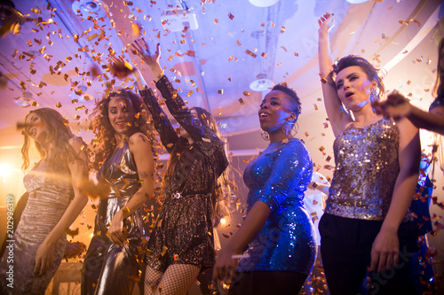Multi-ethnic group of beautiful young women wearing glittering dresses dancing under golden confetti shower enjoying raving party in nightclub