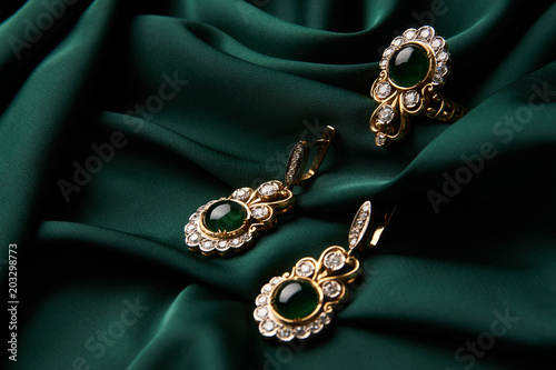 Beautiful Golden ring and pair of earrings with green Emerald and Diamonds gemstones on a green satin background. Luxury female jewelry, close-up. Selective focus