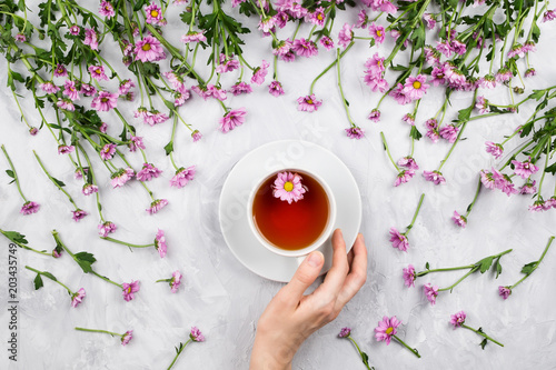 Flatlay with woman's hand holding cup of tea surrounded by pink daisies, cement background