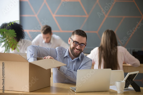 Smiling new male employee unpacking box with belongings at workplace, happy hired office worker newcomer on first working day concept, excited millennial businessman put laptop on desk in coworking