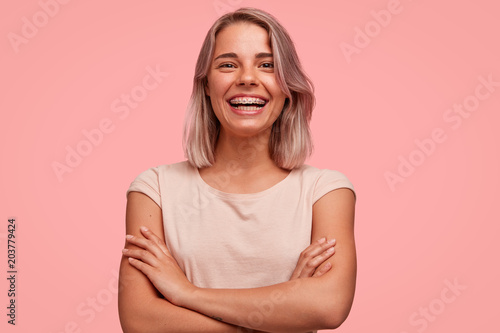 People, happiness and facial expressions concept. Pretty young woman with broad shining smile, keeps hands crossed, being in high spirit, wears braces on teeth, poses alone against pink background