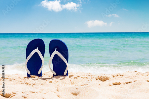Pair of flip flops on sand beach with tropical blue sea and sky in background in summer 