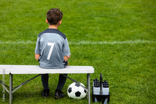 Little footballer sitting on a wooden bench and watching soccer game. Young substitute player waiting on a soccer bench. Boy on a grass pitch with a soccer ball and water bottles.