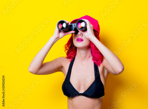 Young pink hair girl in bikini with cap and binoculars. Portrait isolated on yellow background