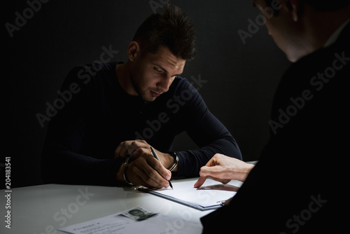 Criminal man with handcuffs signing document in interrogation room