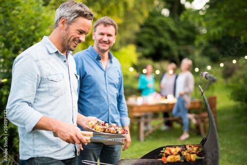 in a summer evening, two men in their forties prepares a barbecue for friends gathered around a table in the garden