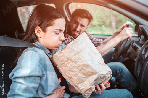 A picture of sick girl holding a bag and trying to vomit into it. She feels bad. Girl is holding her hand on stomach. Guy is looking at her with sight full of disgution.