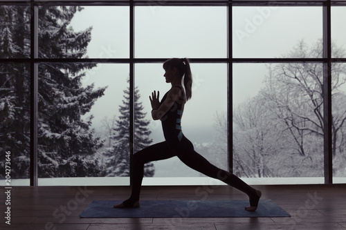 A silhouette of a woman doing yoga on background of windows with beautiful winter landscape with trees in the snow and sunlight, young yogi girl in warrior's yoga position