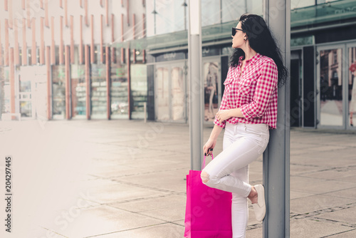 The woman is back from shopping. A girl wearing white jeans and a checkered shirt is holding shopping bags in her hands staring at the shopping center.
