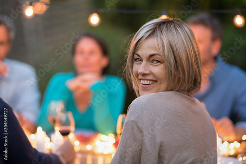 One summer evening, friends gathered around a table in the garden lit by light garlands. Focus on a pretty woman