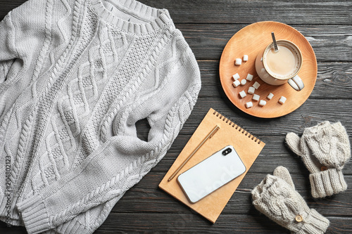 Flat lay composition with warm knitted clothes, smartphone and cup of cocoa on wooden background