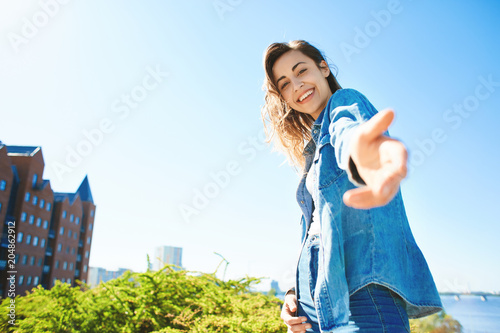 portrait of a young smiling attractive woman in jeans clothes at sunny day on the blue sky background. happy woman gives a hand to someone like follow me . first person view.