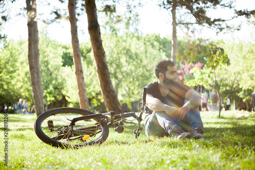 Young bearded man sitting on the ground next to his bicycle in the park. The sun rays are crossing through the trees