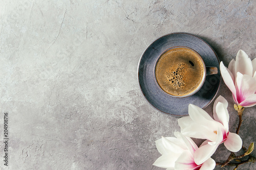 Blue cup of black espresso coffee and spring flowers magnolia branches over grey texture background. Top view, copy space. Spring greeting card, wallpapers