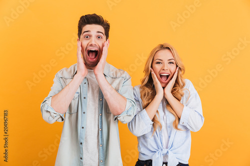 Image of ecstatic people man and woman in basic clothing screaming in surprise or delight and touching cheeks, isolated over yellow background